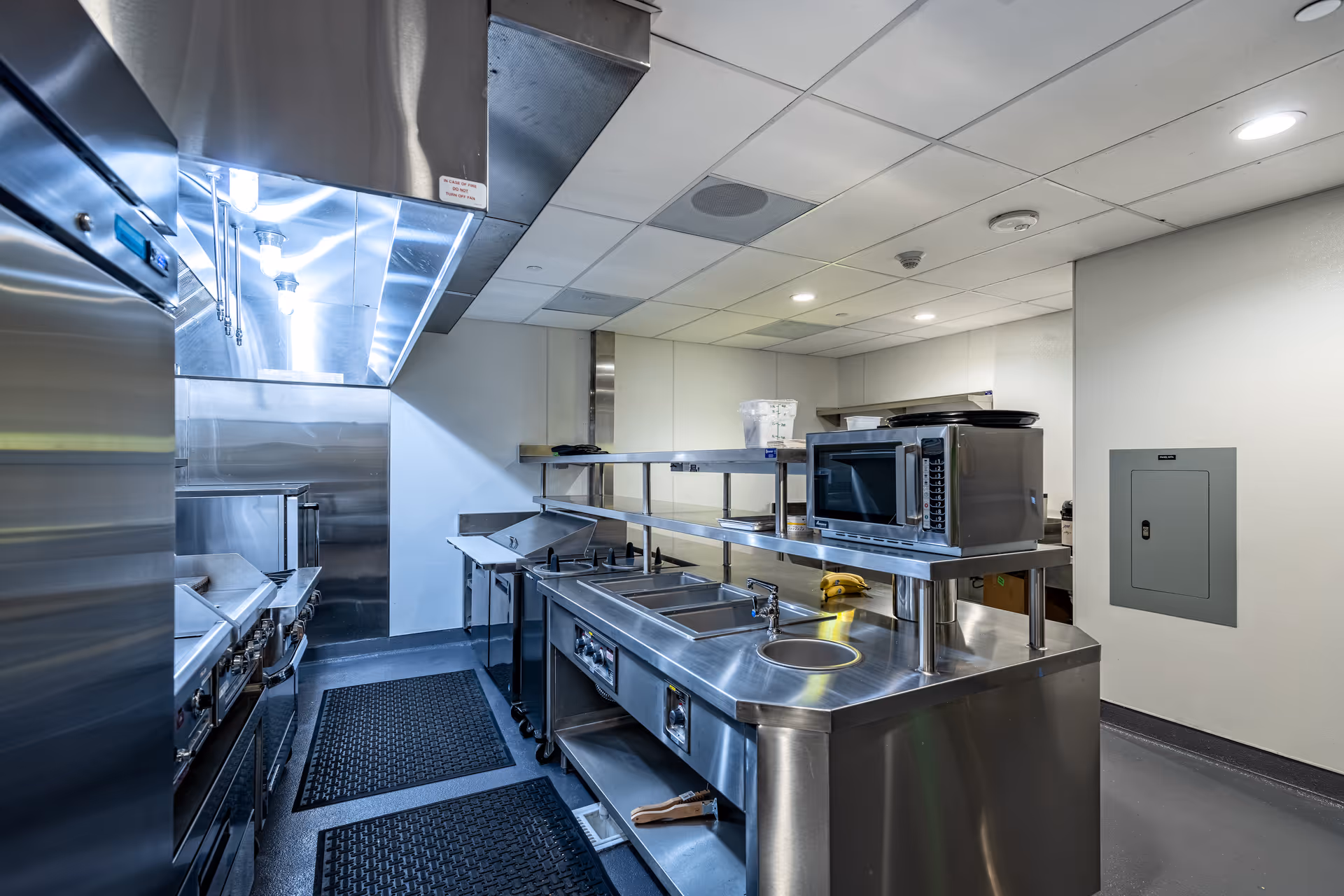 A clean, modern commercial kitchen with stainless steel appliances and countertops, including a microwave, stove, and sink. The room has a white ceiling with recessed lighting and a gray floor with black mats.