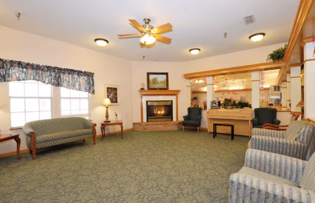 A spacious living room with patterned green carpet, several upholstered chairs and sofas arranged around the room, a ceiling fan with lights, a lit fireplace with a painting above it, and a counter area with plants and decorations in the background.