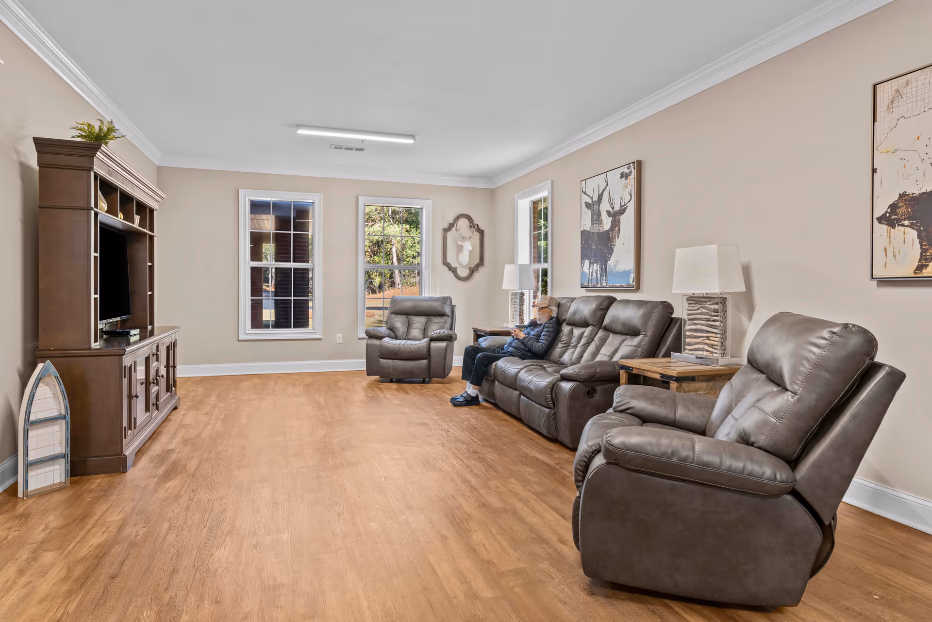 A spacious living room with wooden flooring, beige walls, and three large windows letting in natural light. The room is furnished with a dark brown leather sofa, two matching recliners, a wooden entertainment center with a TV, and two side tables with lamps. A person is seated on the sofa. The walls are decorated with framed animal artwork and a decorative mirror.