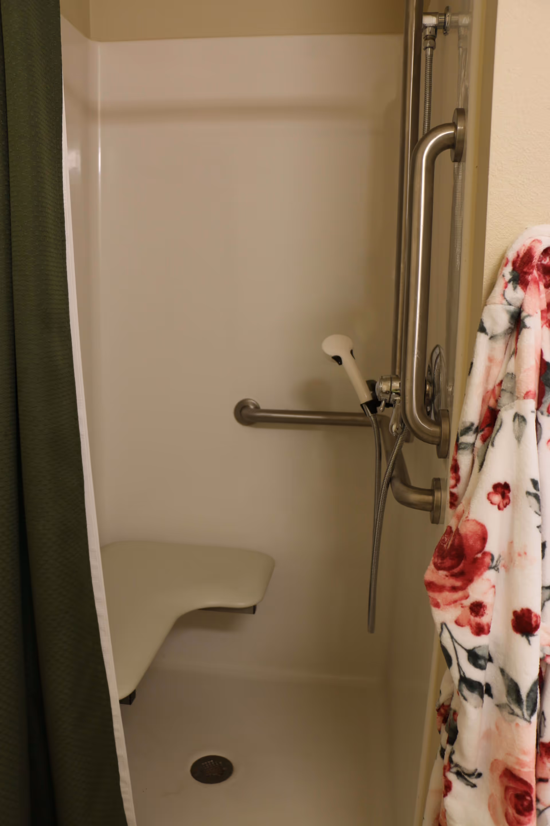 Interior of a senior living facility shower with a fold-down seat, grab bars, a handheld showerhead, a green shower curtain, and a floral patterned bathrobe hanging on the side.