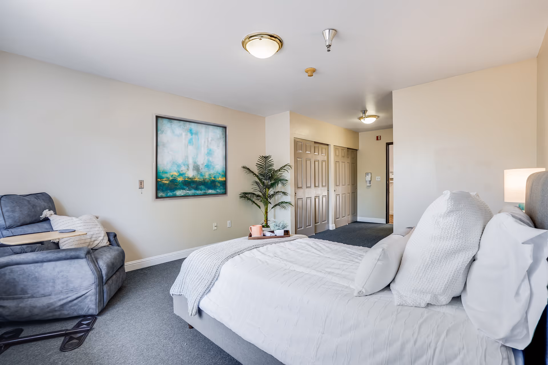 A bright, neatly furnished bedroom in a senior living unit with a bed, reclining armchair, wall art, and double closets.