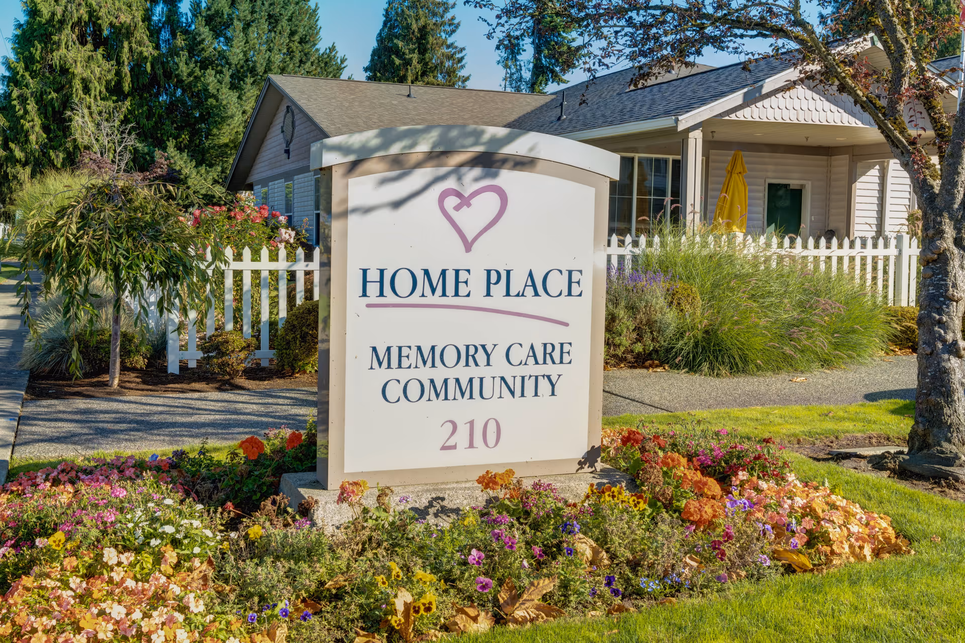 Outdoor view of a sign for Home Place Memory Care Community with the number 210, surrounded by colorful flowers and greenery, with a white picket fence and a building in the background.
