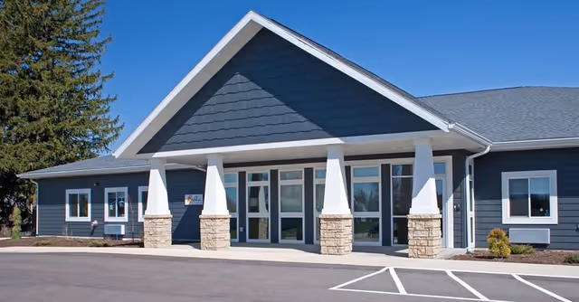 Front exterior view of a single-story building with dark blue siding, white trim, and stone pillars supporting a covered entrance. The building has multiple windows and a clear blue sky above. There is a parking area in front with marked spaces.