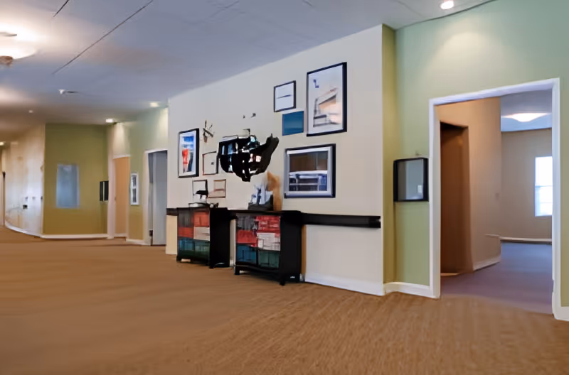 Interior hallway of a senior living facility with beige carpet and light green and cream walls. The hallway features a wall decorated with framed pictures and two colorful storage cabinets. There are open doorways leading to other rooms along the hallway.