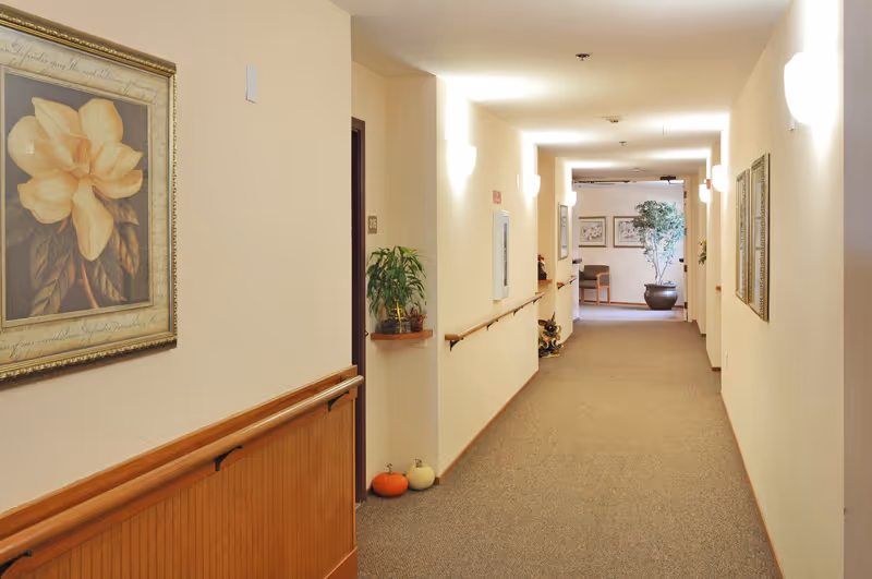 Well-lit carpeted hallway in a senior living facility with handrails, framed artwork, small pumpkins and potted plants leading to a seating area at the far end.