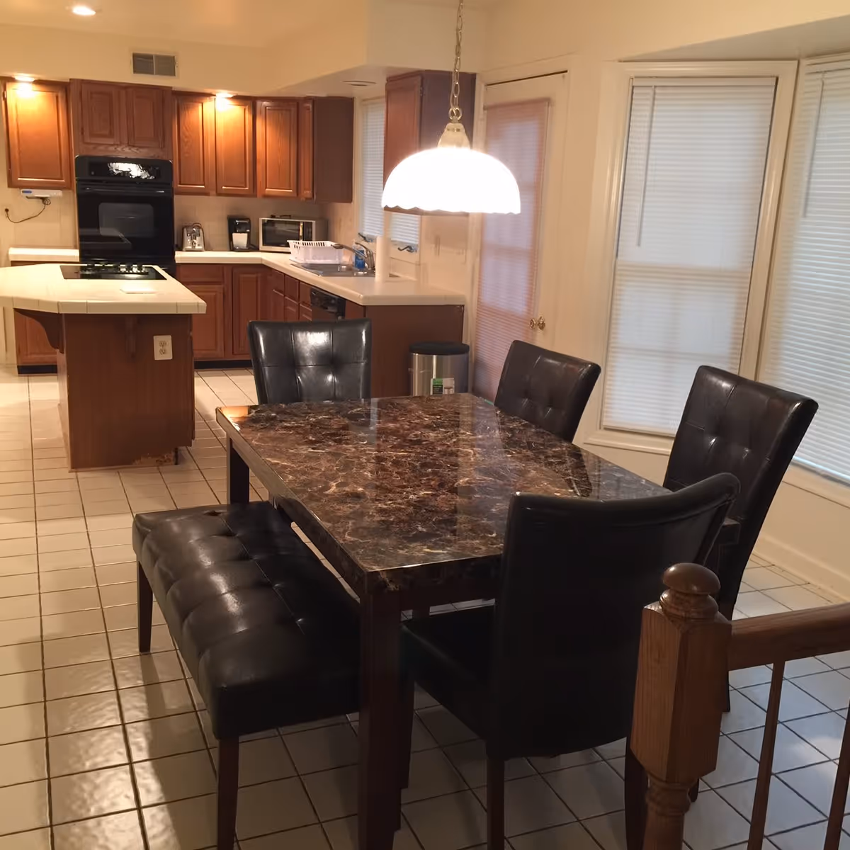 A kitchen and dining area featuring a dark marble dining table with four black leather chairs and a matching bench. The kitchen has wooden cabinets, a white tiled countertop with a built-in stove, an oven, a microwave, and a coffee maker. A hanging light fixture illuminates the dining table, and windows with blinds are visible in the background.