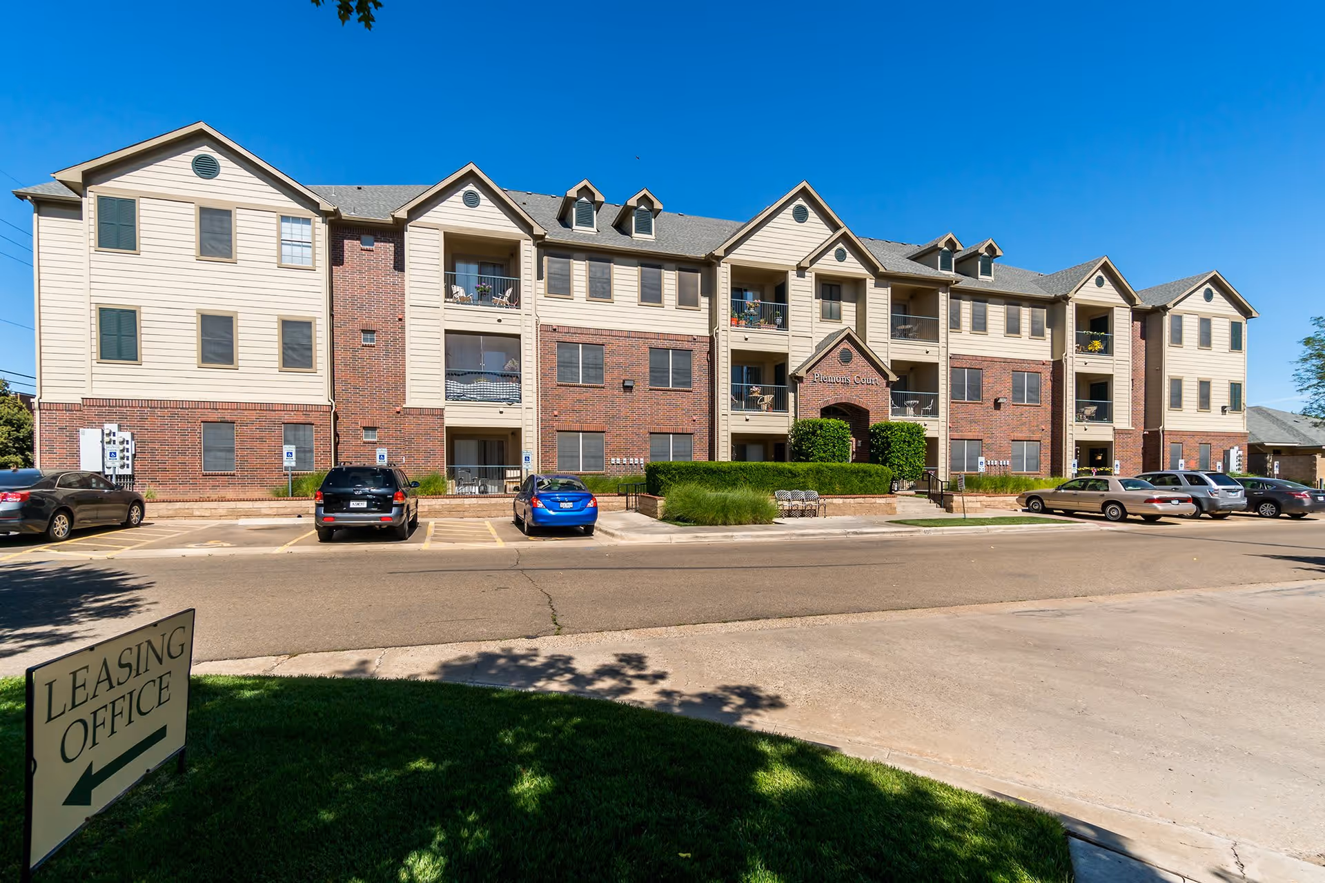 Exterior view of a three-story residential building with beige siding and red brick accents under a clear blue sky. Several cars are parked in front of the building, and a sign on the grass points to the leasing office. The building has balconies with chairs and plants, and a central entrance labeled 'Plemons Court'.