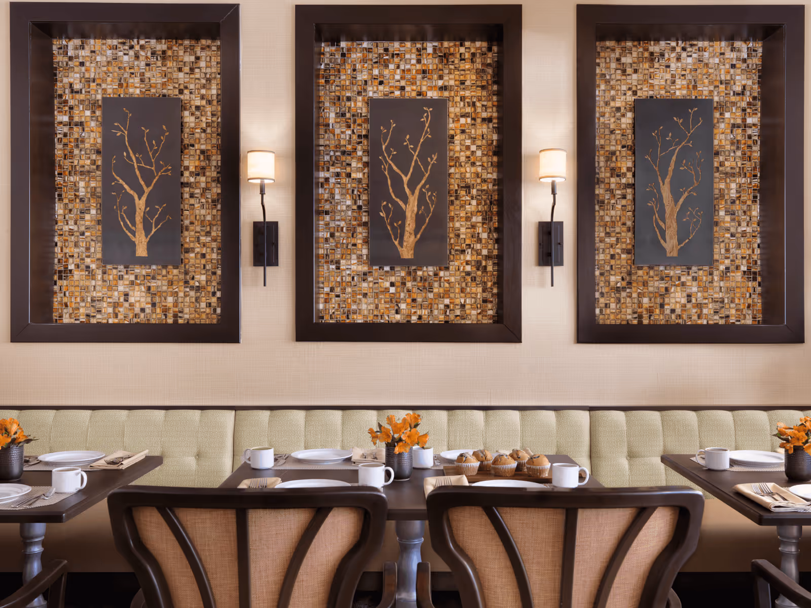 A dining area with a long cushioned bench along the wall, dark wooden tables set with white plates, cups, napkins, and silverware. There are small vases with orange flowers on the tables. The wall behind features three framed artworks of tree branches on a mosaic tile background, each illuminated by a wall sconce with a beige lampshade.