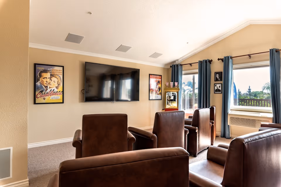 A cozy senior living common area with four brown leather armchairs facing a large flat-screen TV mounted on a beige wall. The room has two large windows with blue curtains, allowing natural light to brighten the space. On the wall are framed vintage movie posters, and a popcorn machine is placed near the windows.