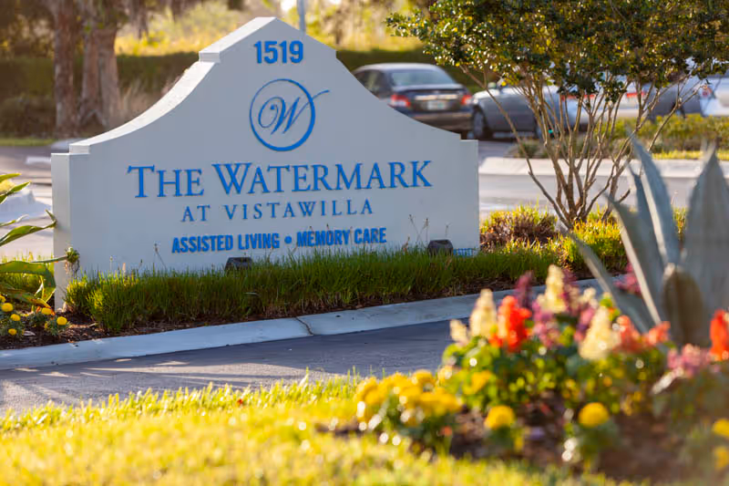 Entrance sign reading "The Watermark at Vistawilla — Assisted Living • Memory Care" surrounded by landscaping and flowers with parked cars in the background.