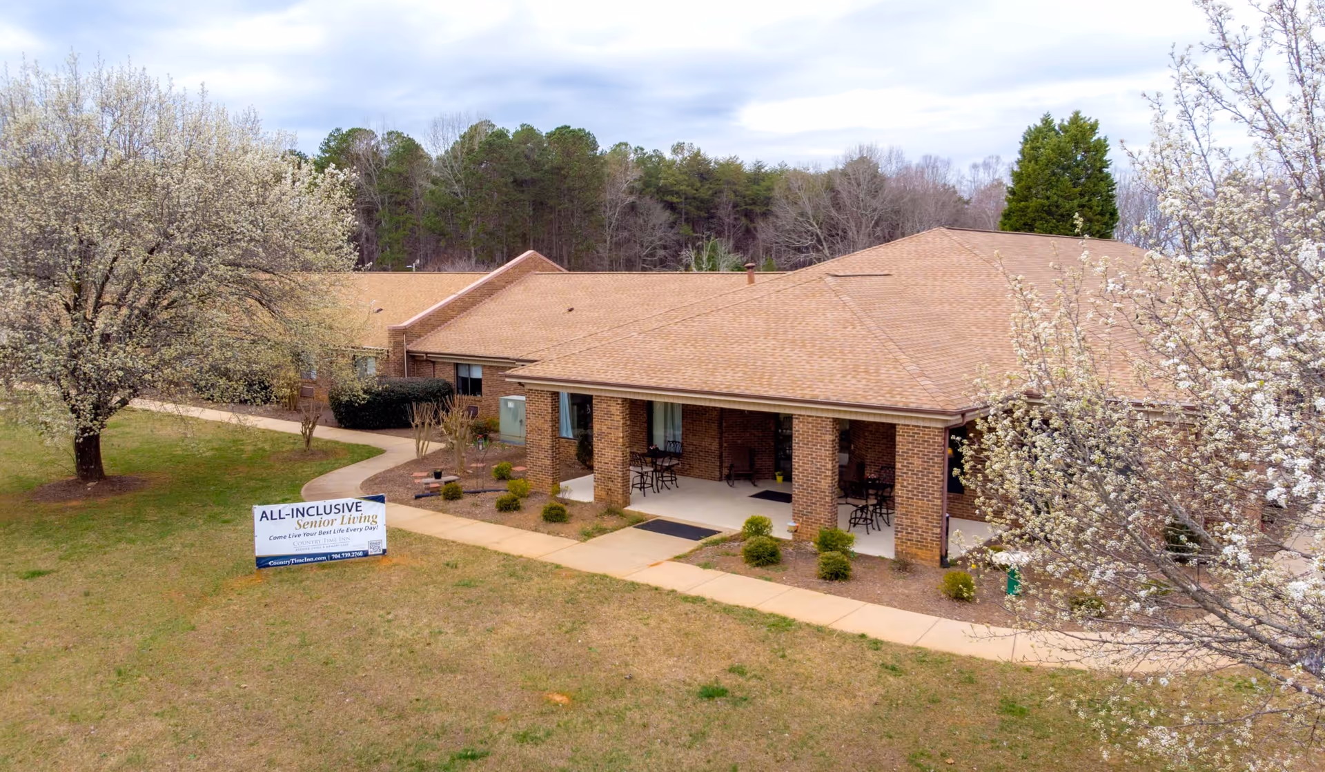 Exterior view of a single-story brick senior living facility with a covered porch featuring several chairs and small tables. The building is surrounded by a grassy area with blooming trees and a paved walkway leading to the entrance. A sign on the lawn reads 'ALL-INCLUSIVE Senior Living Come Live Your Best Life Every Day.'