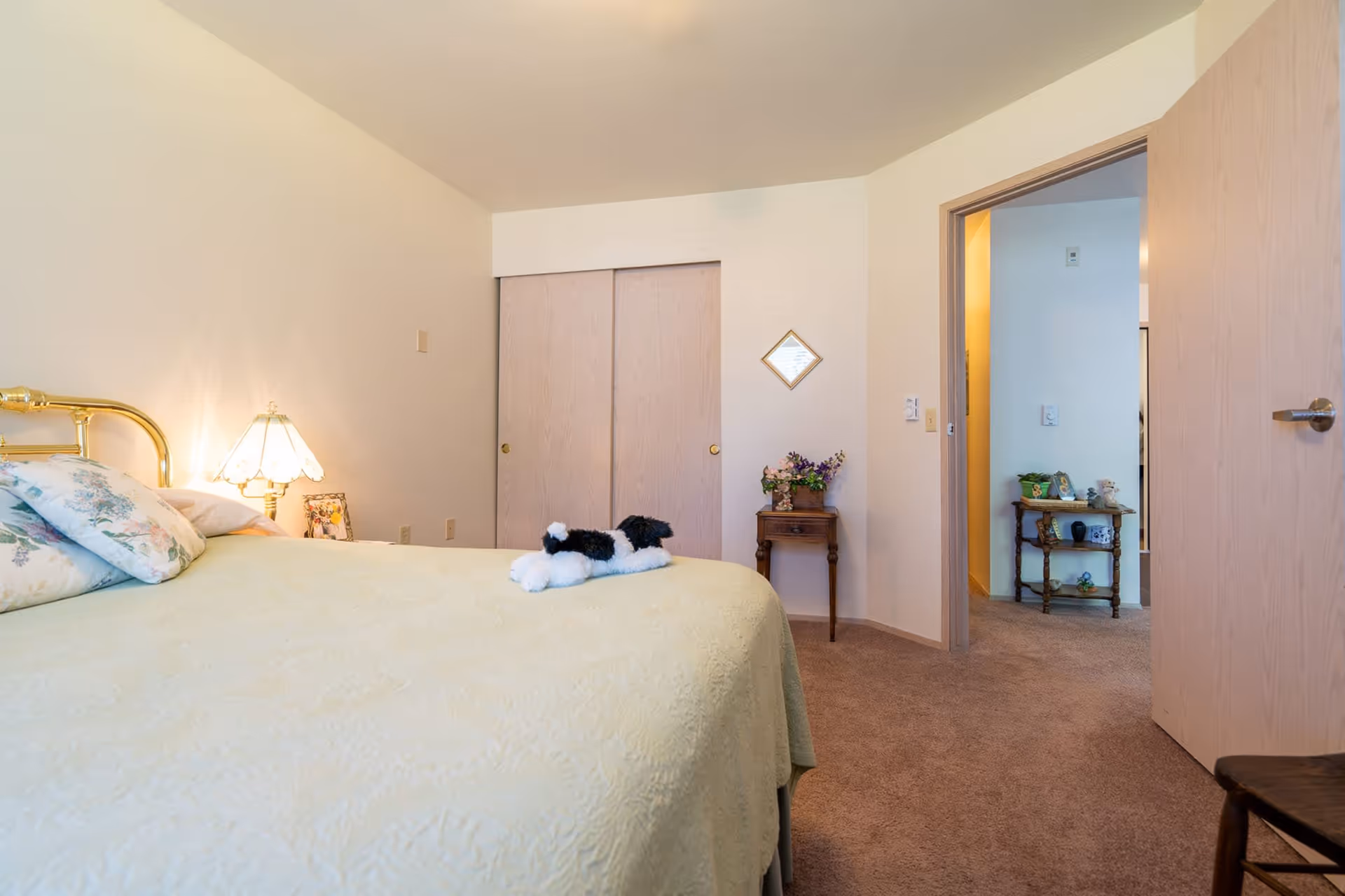 A bedroom with a bed covered in a light yellow quilt and floral pillows. A small stuffed dog toy is placed on the bed. There is a brass headboard and a bedside lamp with a floral lampshade. The room has beige carpet, a small wooden side table with a flower arrangement, a wall mirror, and a closet with sliding wooden doors. An open door leads to another room with a small wooden shelf holding decorative items.