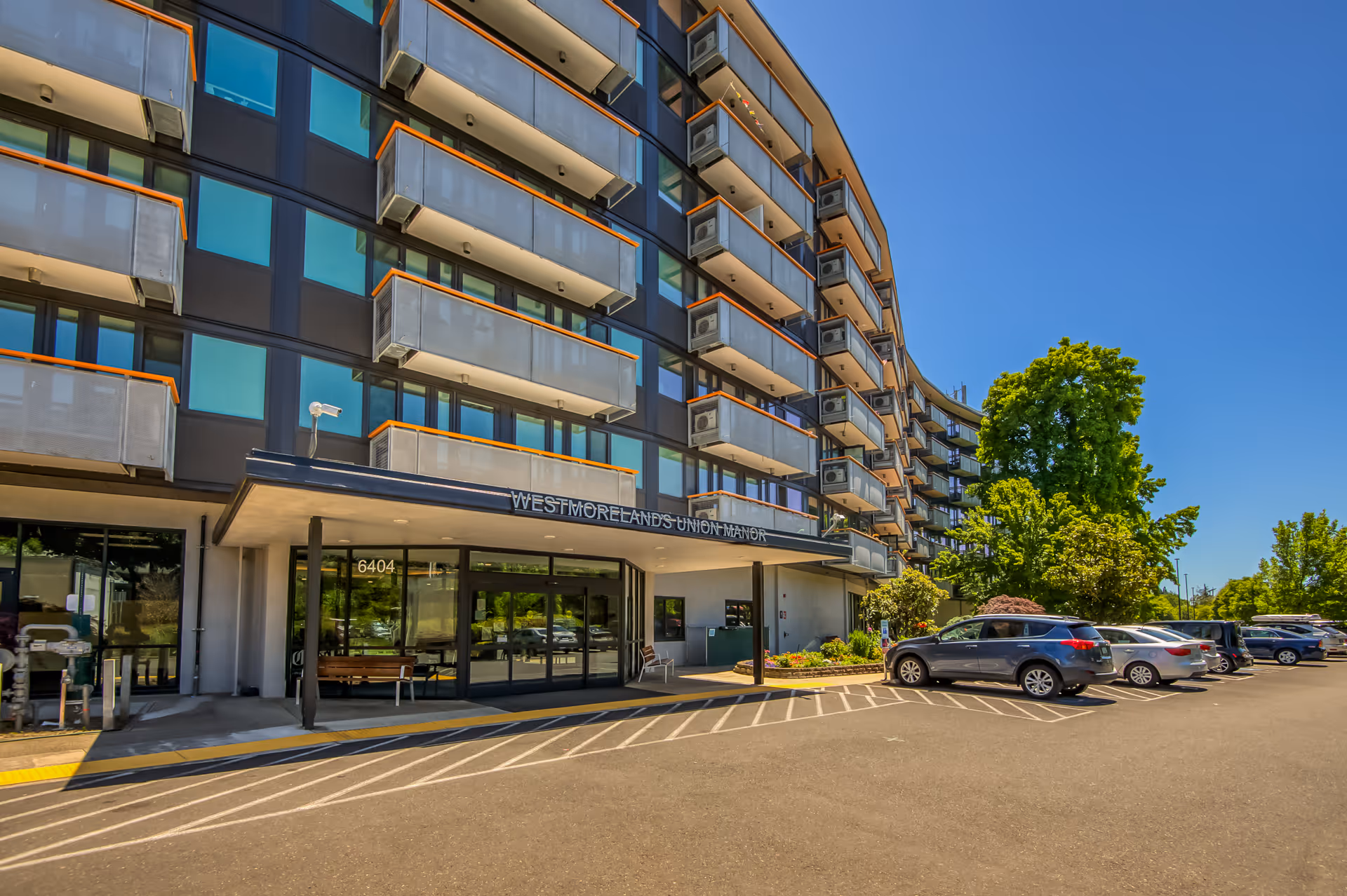 Exterior view of Westmoreland's Union Manor, a multi-story building with balconies and large windows. There are several parked cars in front of the entrance under a canopy with the facility's name displayed. Trees and clear blue sky are visible in the background.