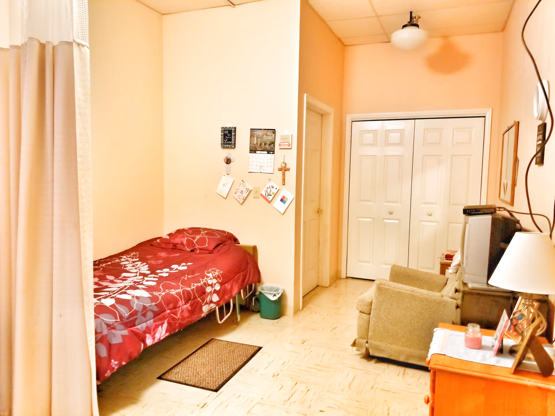 A small, cozy room with a single bed covered in a red floral bedspread, a beige armchair, a wooden dresser with a lamp and framed photos, a TV on a stand, and a closet with white double doors. The walls are light peach, and there is a curtain partially visible on the left side.
