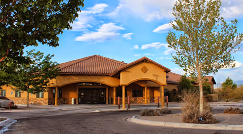 Exterior view of a single-story assisted living facility building with a tiled roof, arched entrance, and surrounding trees under a partly cloudy sky.