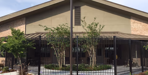 Brick nursing facility front with black metal fence and trees.