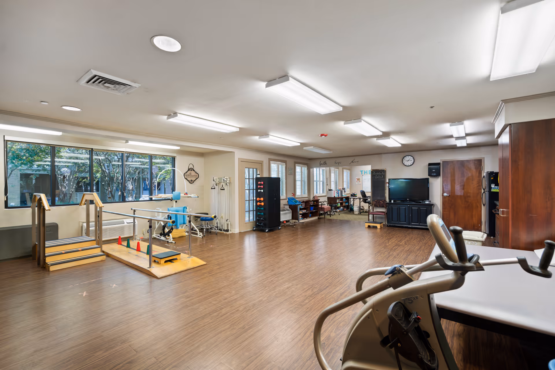 A spacious rehabilitation or physical therapy room with wooden flooring, exercise equipment including a stationary bike, parallel bars with stairs, dumbbells on a rack, and a TV mounted on a cabinet. Large windows let in natural light, and there are chairs and tables in the background near a clock on the wall.