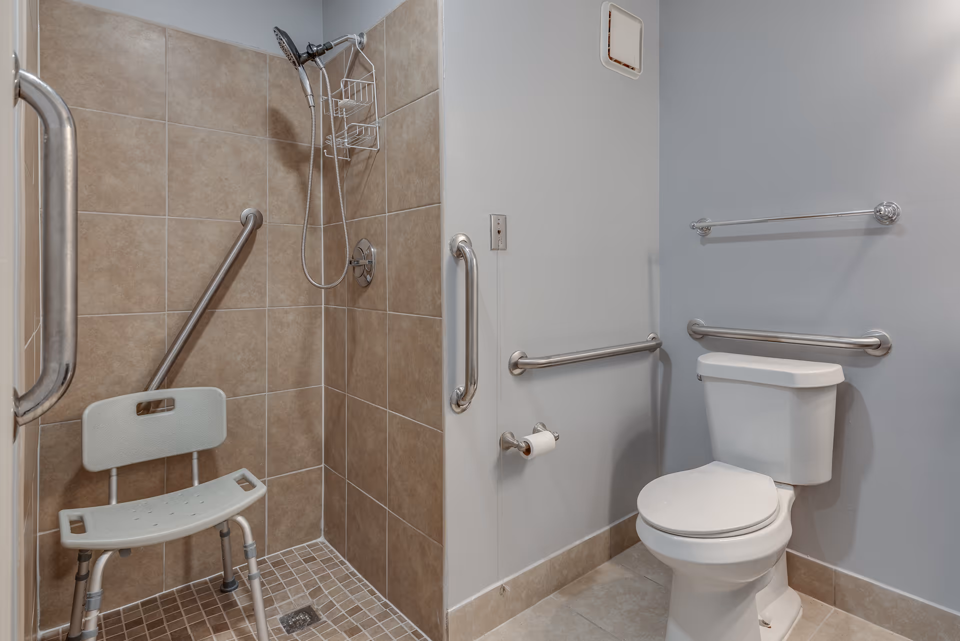 Accessible bathroom with a tiled walk-in shower featuring a handheld showerhead and a shower chair. The bathroom also includes a toilet with grab bars on the walls for support and safety.