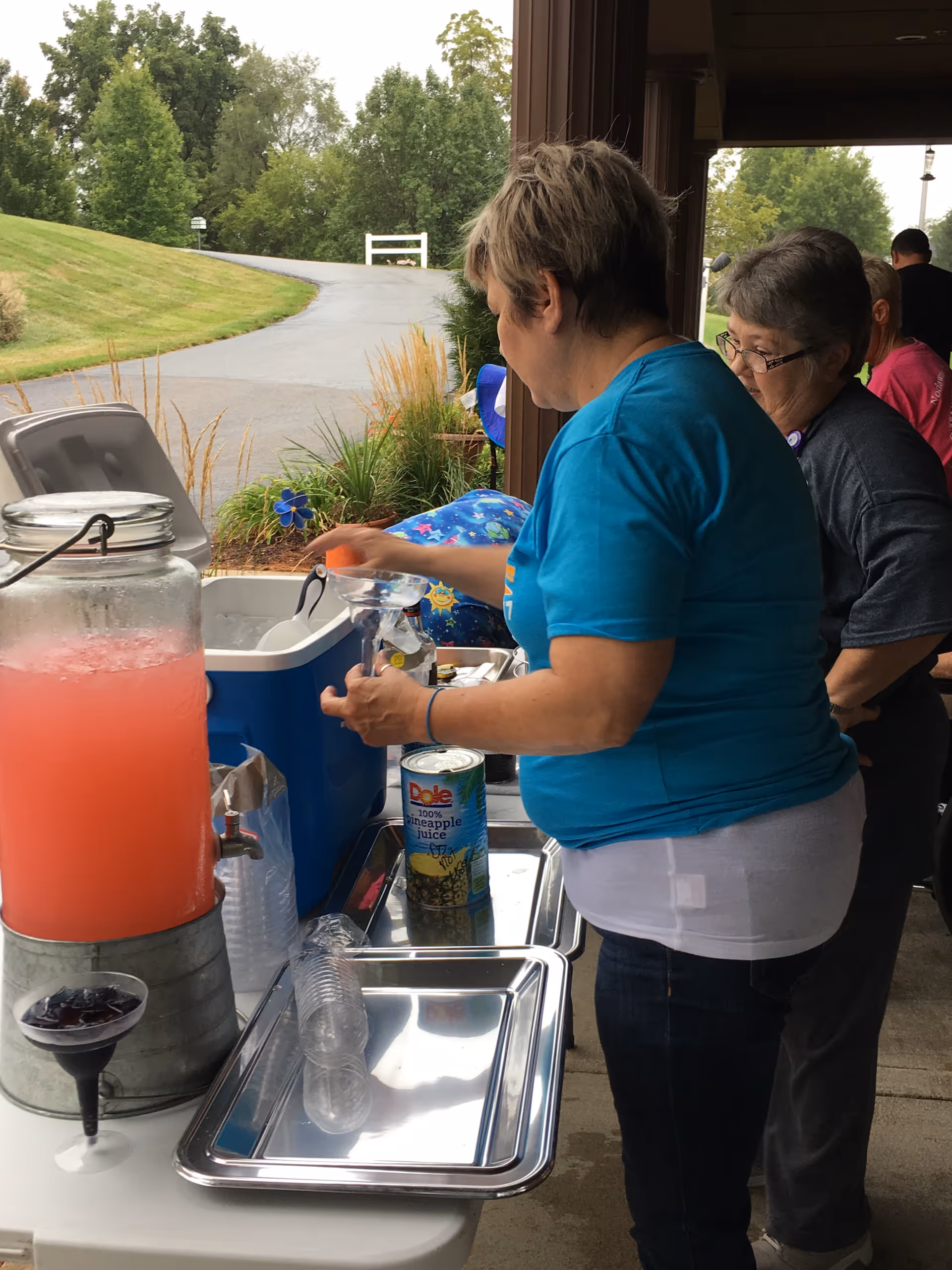 Two women standing at a table outdoors near a building, one woman is pouring a drink from a cooler while the other watches. On the table are a large container of pink beverage, a can of Dole pineapple juice, plastic cups, and trays. The background shows a paved path, grass, and trees.