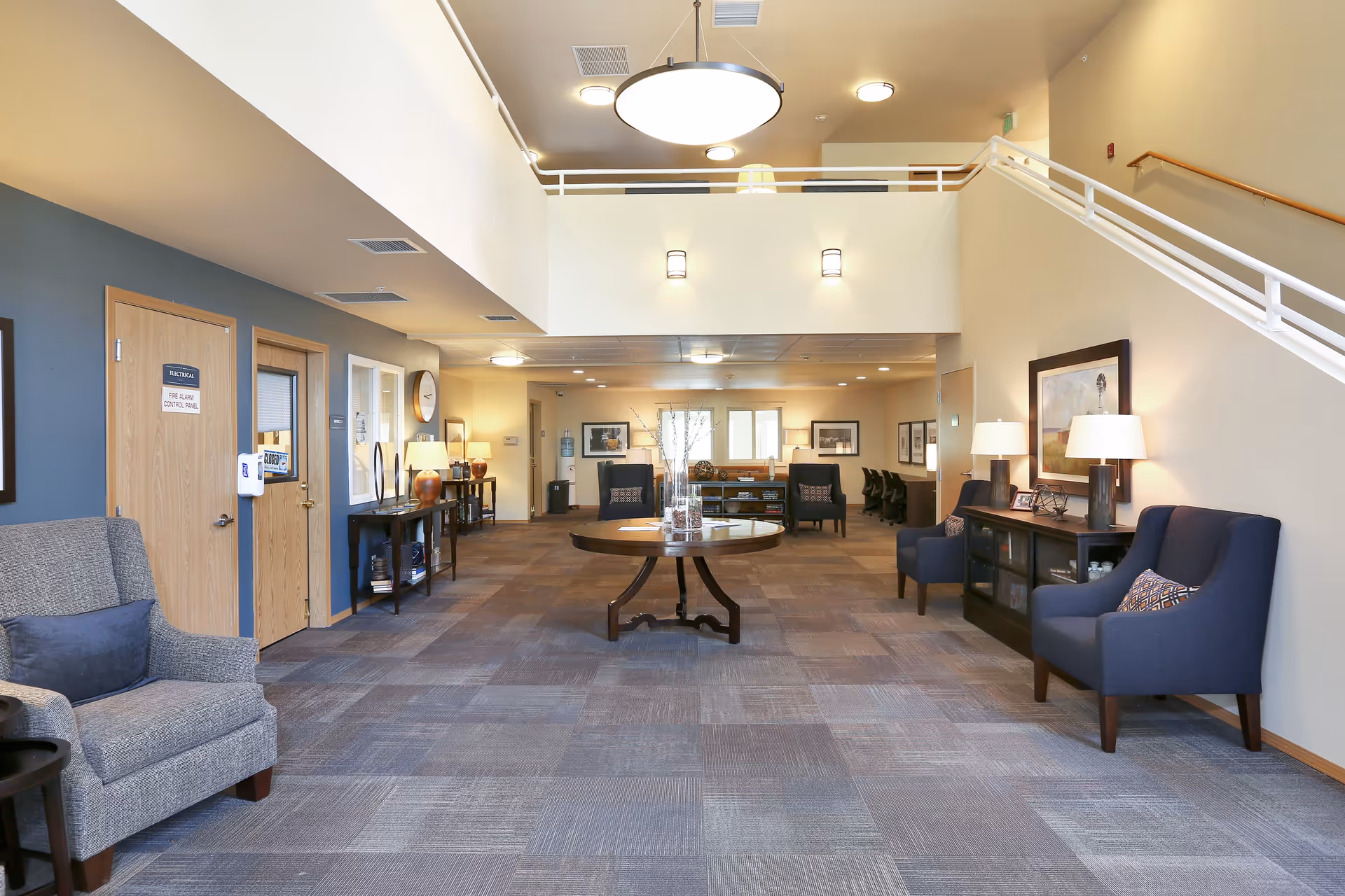 Spacious senior living facility common area with high ceilings, carpeted floor, and modern lighting. The room features a round wooden table with decorative items in the center, several upholstered armchairs along the walls, framed artwork, and a staircase with white railings leading to an upper level. The walls are painted in neutral tones with a blue accent wall on the left side.