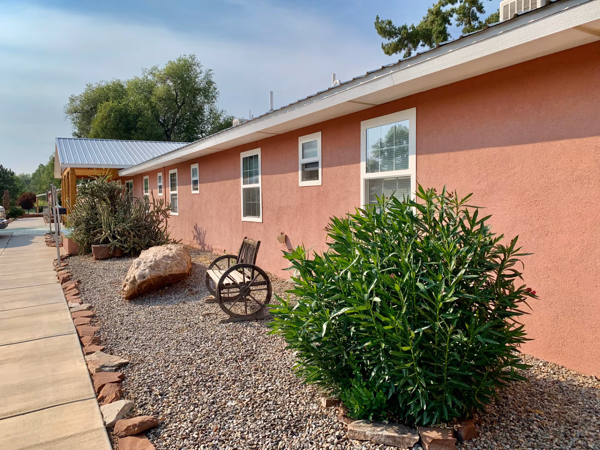 Exterior view of a single-story building with a pink stucco wall and several windows. In front of the building, there is a landscaped area with gravel, a large rock, a wooden bench with wagon wheel sides, and various green plants and bushes. A concrete sidewalk runs alongside the building under a partly cloudy sky.