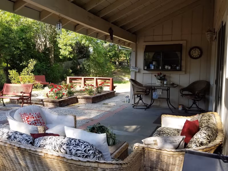 Covered outdoor patio area with wicker furniture including a sofa and chairs with cushions and pillows. A small table with two chairs is against the wall under a window. There are flower beds with red flowers and benches in the garden area beyond the patio, surrounded by trees and greenery.