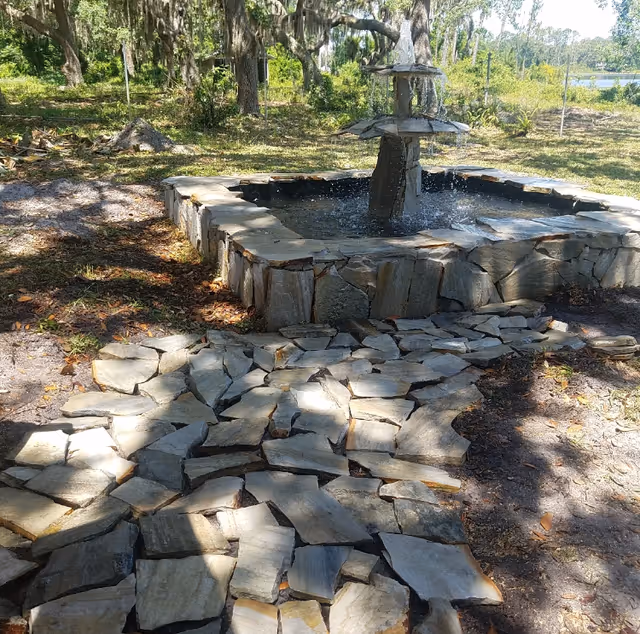 Outdoor stone water fountain with water flowing, surrounded by a stone pathway and trees in the background under sunlight.