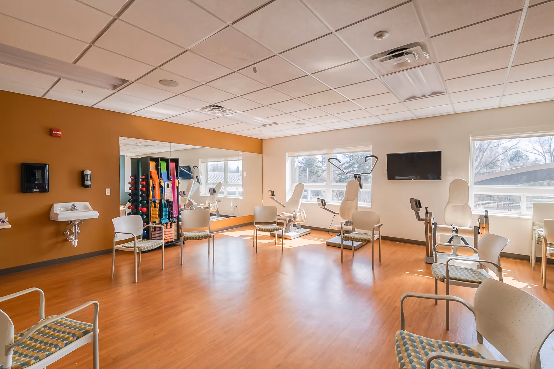 A bright exercise room with wooden flooring, several chairs arranged in a circle, exercise equipment including stationary bikes and resistance bands, a wall-mounted TV, large windows letting in natural light, and a wall mirror reflecting part of the room.