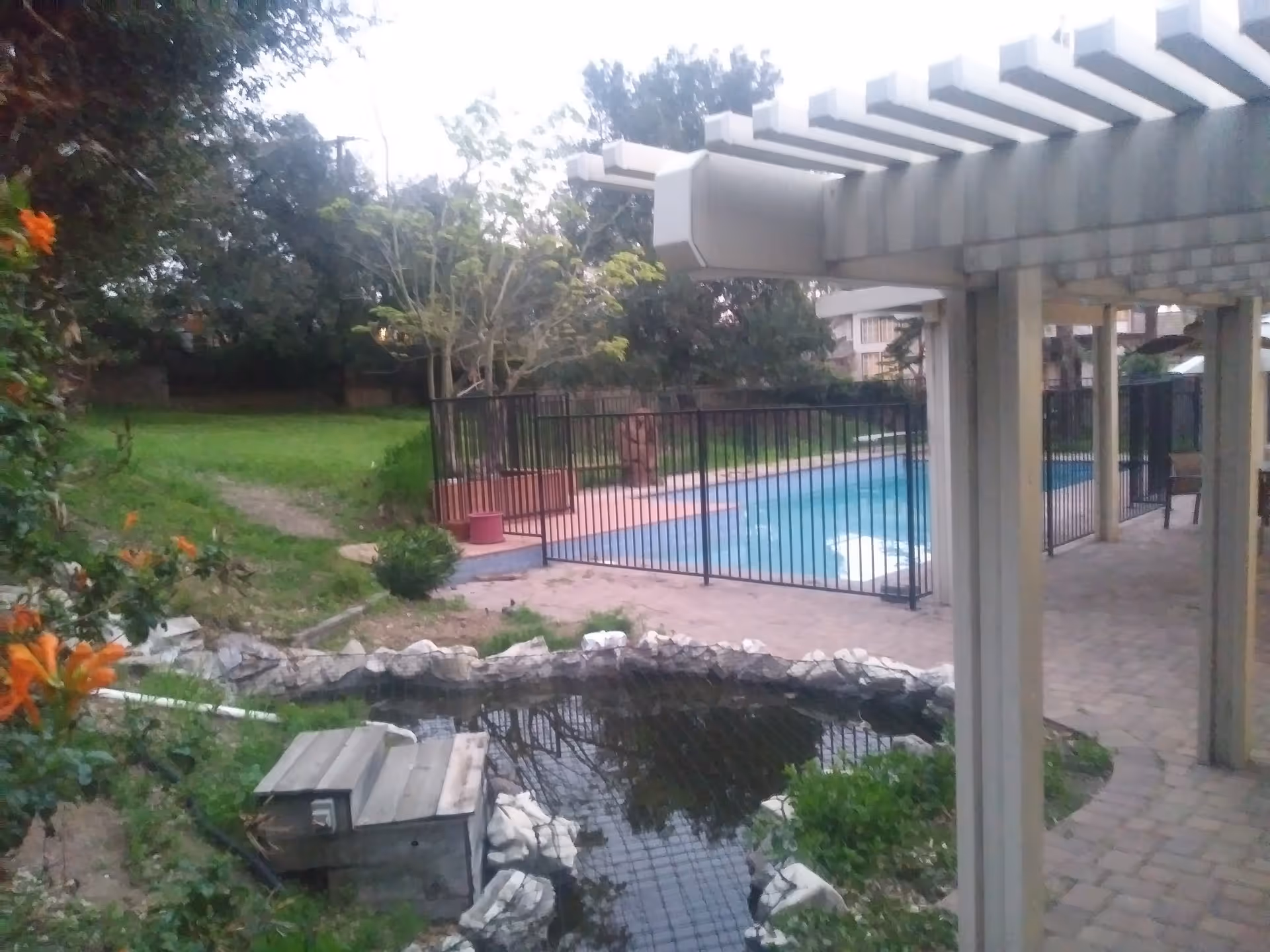 Outdoor area featuring a small pond with rocks around it, a wooden structure beside the pond, a fenced swimming pool, green grass, trees, and a covered patio with columns and a pergola-style roof.