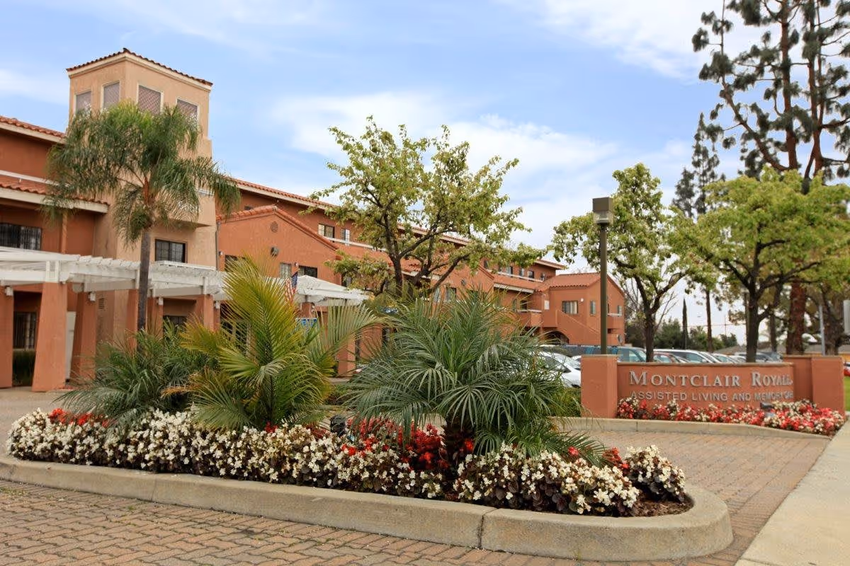 Exterior view of Montclair Royale assisted living and memory care facility with a landscaped garden featuring palm trees and flowering plants in front of the building under a partly cloudy sky.