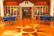 Bright and spacious dining area with polished wooden floor featuring a large sunburst design. Two round tables with blue tablecloths and wooden chairs are set near open double doors leading to another room. Flower arrangements are placed on the tables, and the room has large windows with white frames and decorative orange awnings.