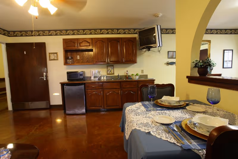 Dining area with a table set with plates and glasses in front of a kitchenette with wooden cabinets, mini fridge and a wall-mounted TV.