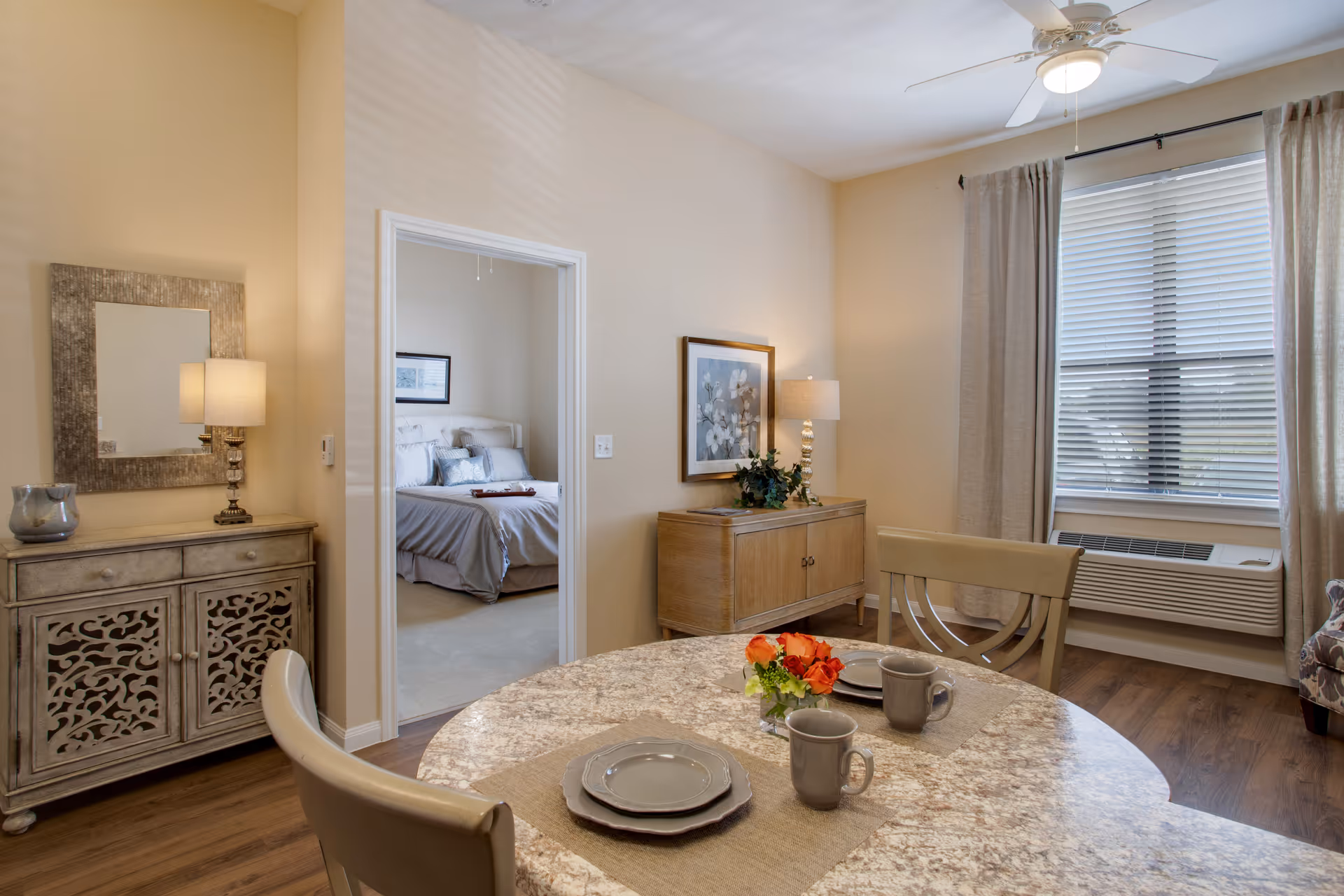 Interior view of a senior living facility showing a dining area with a round table set with two plates, two mugs, and a small flower arrangement. The room has wooden flooring, beige walls, a window with blinds and curtains, and a ceiling fan with a light. To the left, there is a doorway leading to a bedroom with a bed and pillows. The dining area also features a sideboard with a lamp and framed artwork above it, and a decorative cabinet with a mirror and lamp on top.