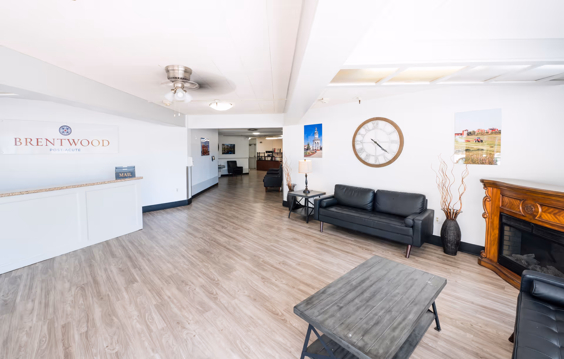 Bright reception lobby with a front desk, seating area with black sofas, wall clock and fireplace at Brentwood Post-Acute.