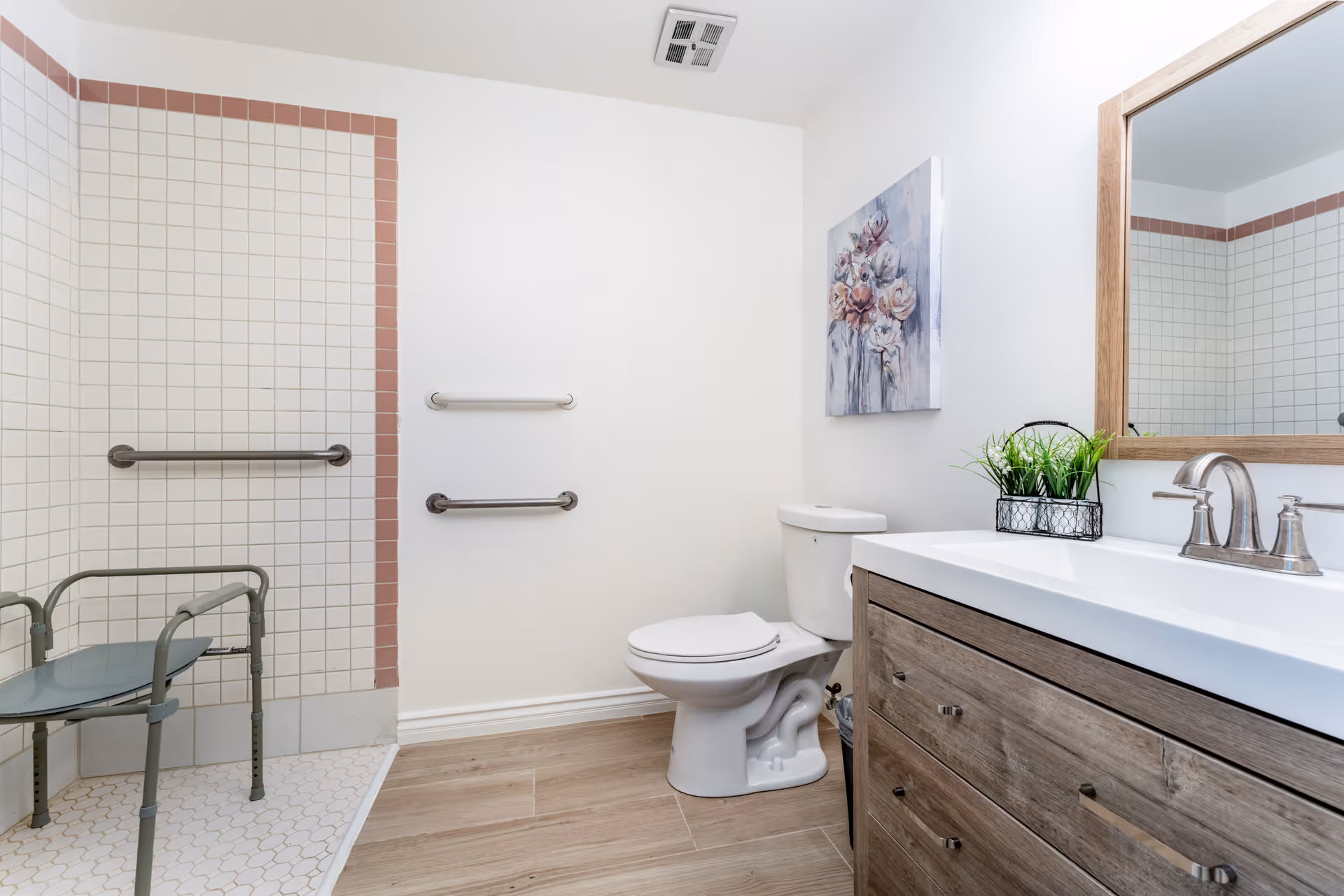 A clean and accessible bathroom with a walk-in tiled shower featuring grab bars and a shower chair. The bathroom also includes a toilet, a wooden vanity with a white countertop and a silver faucet, a large mirror, a small plant decoration, and a floral painting on the wall.