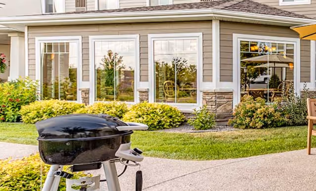 A small black outdoor grill on a patio in front of a senior living building with large windows and landscaped shrubs.