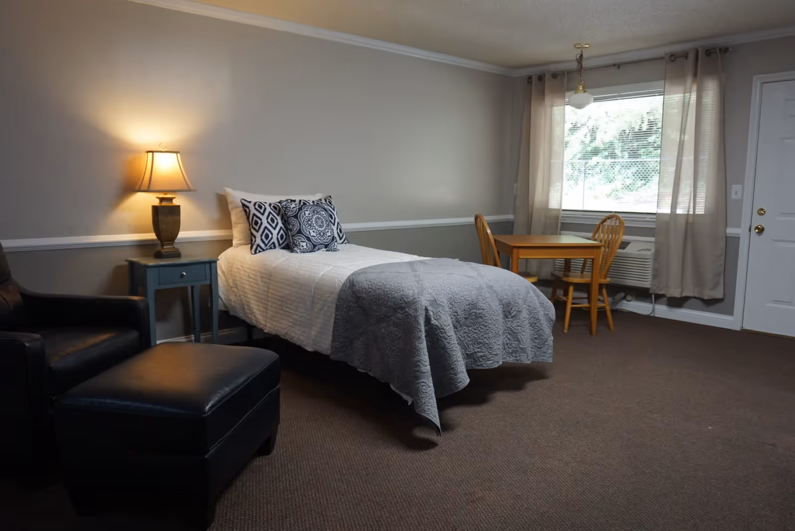 Furnished bedroom with a single bed, nightstand and lamp, armchair with ottoman, and a small table and chairs by a window.