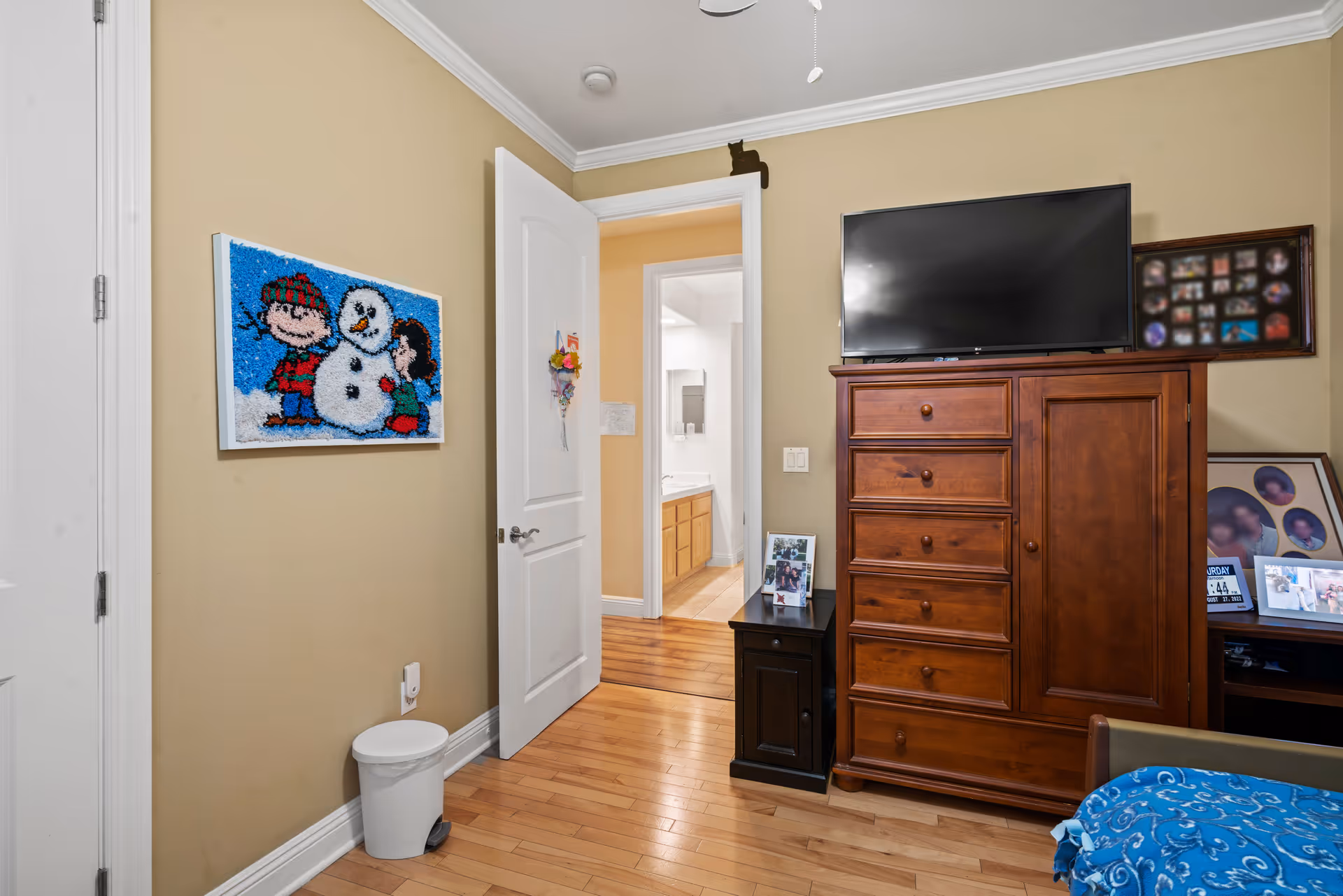 A bedroom with beige walls and hardwood floors featuring a wooden dresser with a flat-screen TV on top, a small black nightstand with framed photos, and a bed partially visible with a blue patterned blanket. A door is open, showing a glimpse of a bathroom with a sink and mirror. A colorful wall art of children building a snowman is hung on the left wall, and a small white trash bin is on the floor below it.