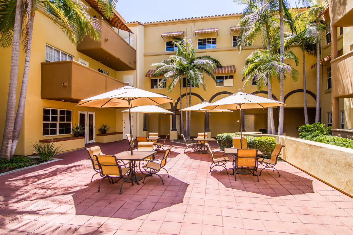 Outdoor courtyard area with several tables and chairs under large beige umbrellas, surrounded by a yellow building with balconies and palm trees providing shade.