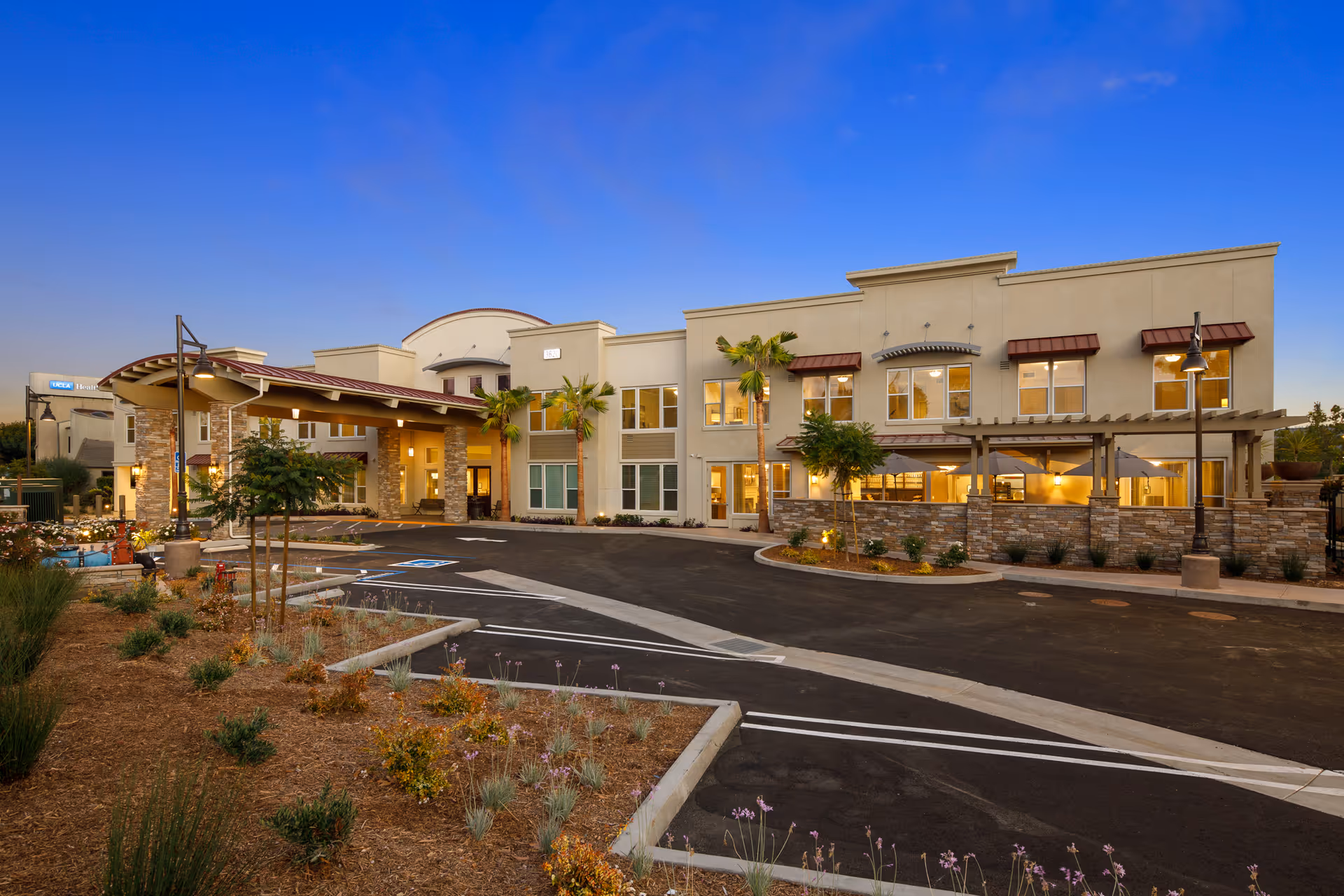 Exterior view of Oakmont of Torrance senior living facility at dusk, showing a two-story building with a covered entrance, palm trees, outdoor seating area with umbrellas, and landscaped surroundings under a clear blue sky.