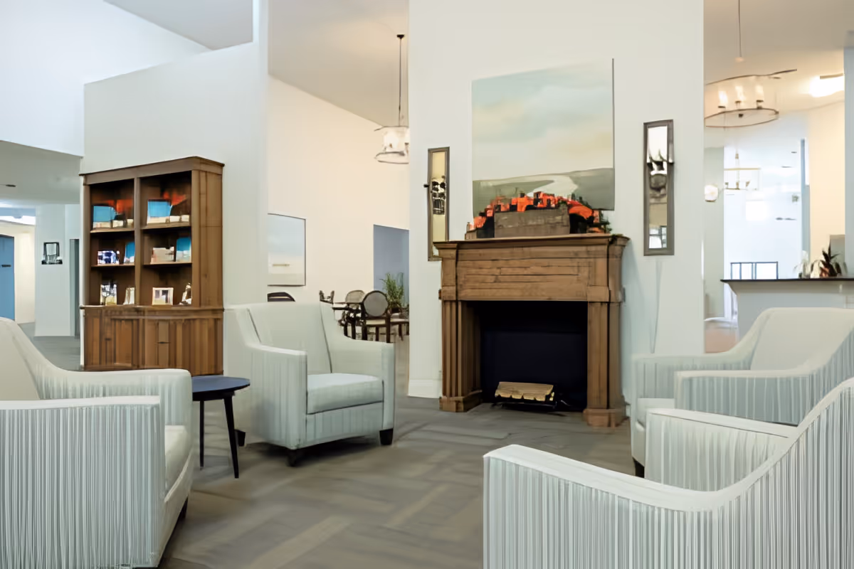 Bright communal lounge with striped armchairs arranged around a wooden fireplace and bookshelf.