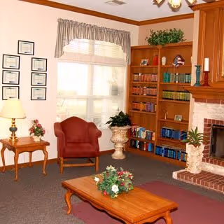 A cozy living room with a red armchair, wooden coffee table with a floral centerpiece, a side table with a lamp and flowers, a large window with curtains, a wooden bookshelf filled with books and decorative items, and a brick fireplace with candles and plants.