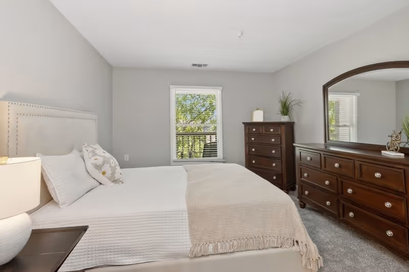 A bedroom with a neatly made bed featuring white bedding and a beige throw blanket. There is a bedside table with a lamp on the left side of the bed. Across from the bed, there is a wooden dresser with a large mirror and another tall wooden chest of drawers. A window with white blinds shows green trees outside. The walls are painted light gray and the floor is carpeted.