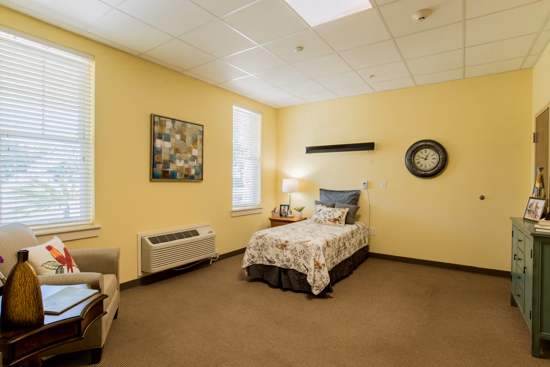 A cozy bedroom in a senior living facility with yellow walls, two windows with white blinds, a single bed with floral bedding, a bedside table with a lamp and framed photos, a wall clock, a green dresser with framed photos, and a comfortable armchair with a decorative pillow and a small table holding a vase and a book.