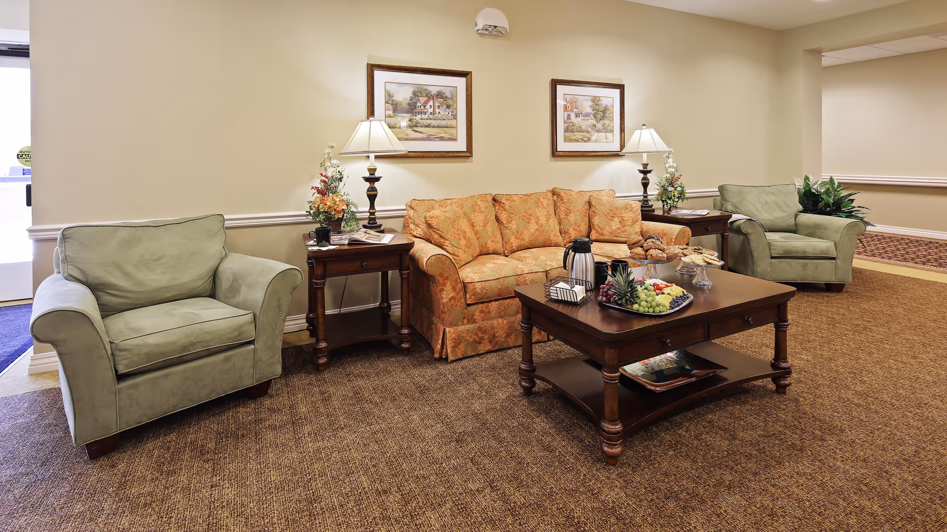 A cozy living room area with a patterned orange sofa flanked by two green armchairs. There are two wooden side tables with lamps and floral arrangements on either side of the sofa. A wooden coffee table in front of the sofa holds a tray with fruit, a coffee pot, and pastries. Two framed paintings hang on the beige wall above the sofa. The room has brown carpet flooring and soft lighting.