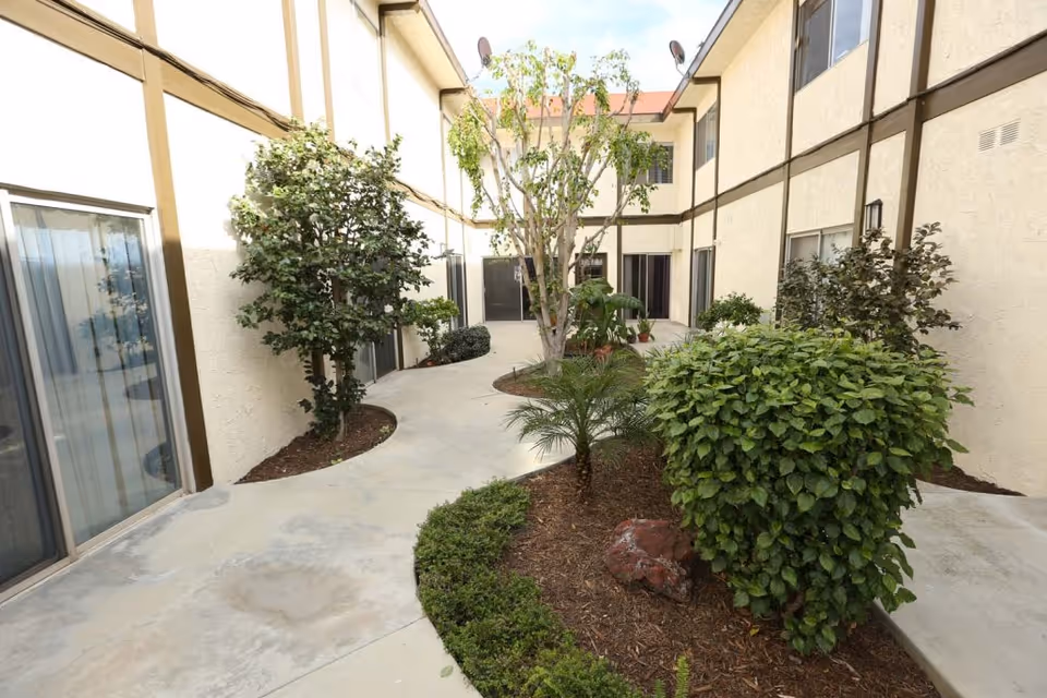 Outdoor courtyard area of Whittier Glen Assisted Living featuring a concrete walkway surrounded by various green shrubs and small trees, with beige building walls and windows on both sides.