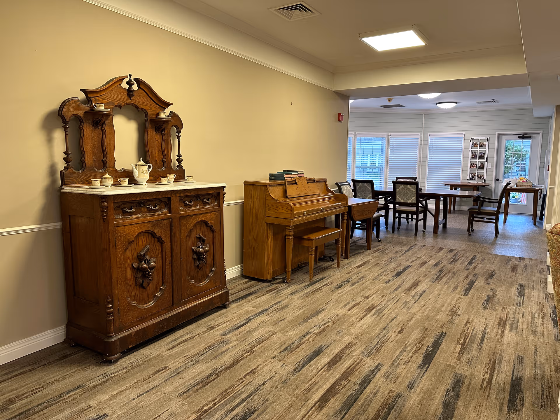A communal interior room showing an antique wooden sideboard and an upright piano along the left wall, with tables and chairs in a dining area beyond.