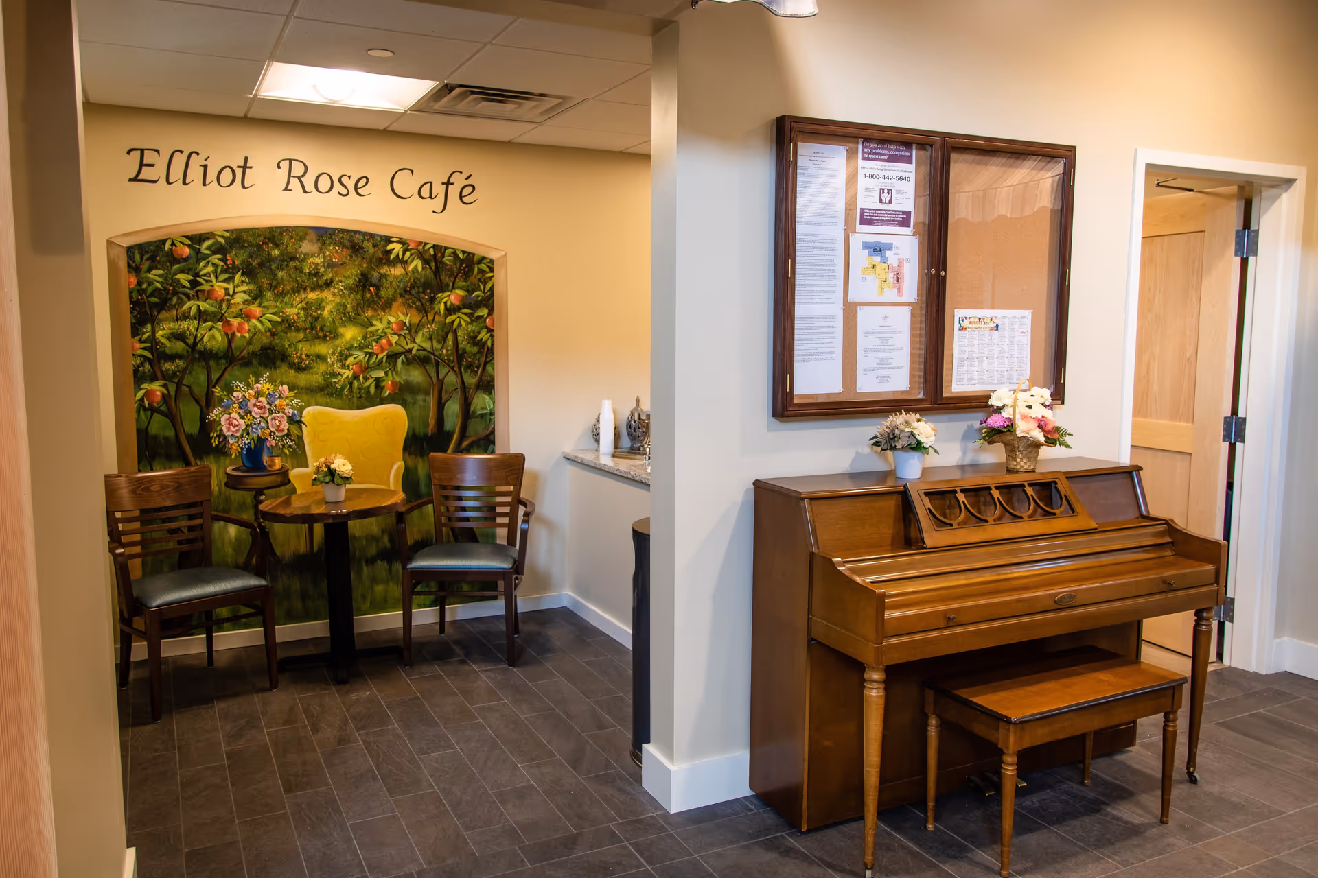 Interior common area with an 'Elliot Rose Café' seating nook featuring a mural and chairs, and a wooden piano with a bench against the wall.