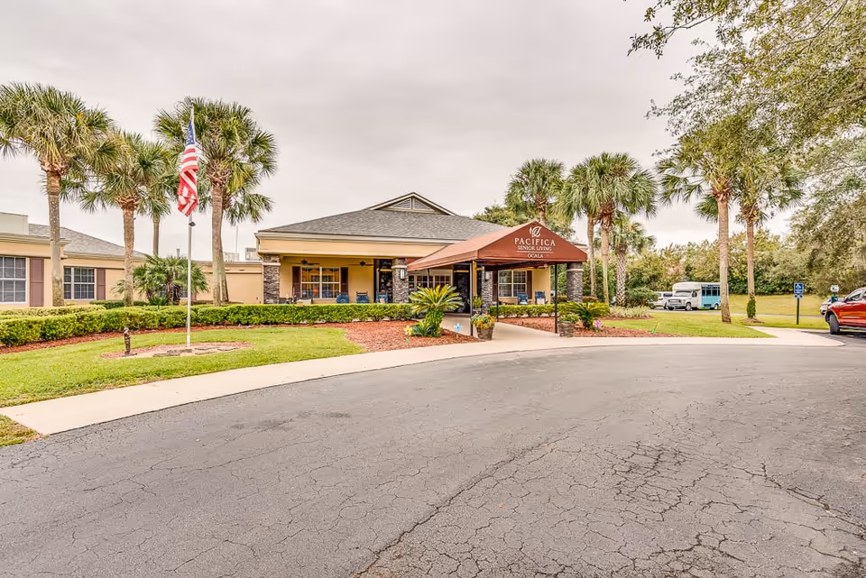 Front exterior view of a senior living facility with a covered entrance canopy labeled Pacifica Senior Living Ocala. The building is surrounded by palm trees, green lawns, and a circular driveway with an American flag on a flagpole.