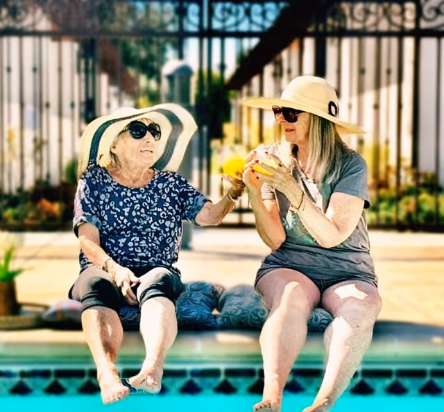 Two elderly women wearing sun hats and sunglasses sitting by the edge of a swimming pool, enjoying drinks and smiling at each other on a sunny day.
